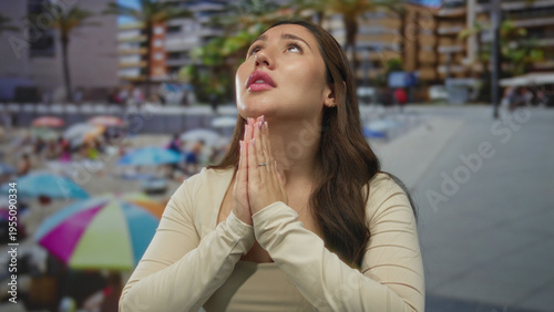 Φωτογραφία Young woman wearing long sleeve top holds hands in prayer and gazes upward amid palm trees and umbrellas at beach; serenity