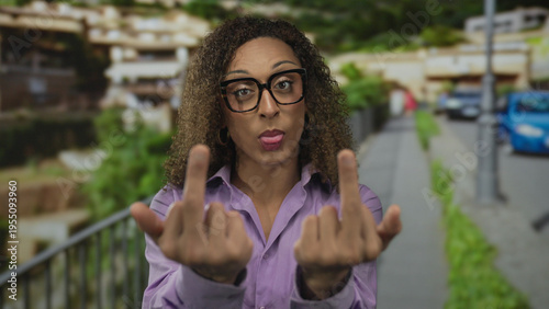 Fotografija Trans woman raising middle finger at camera on street near sidewalk railing amid blurred city buildings; defiance