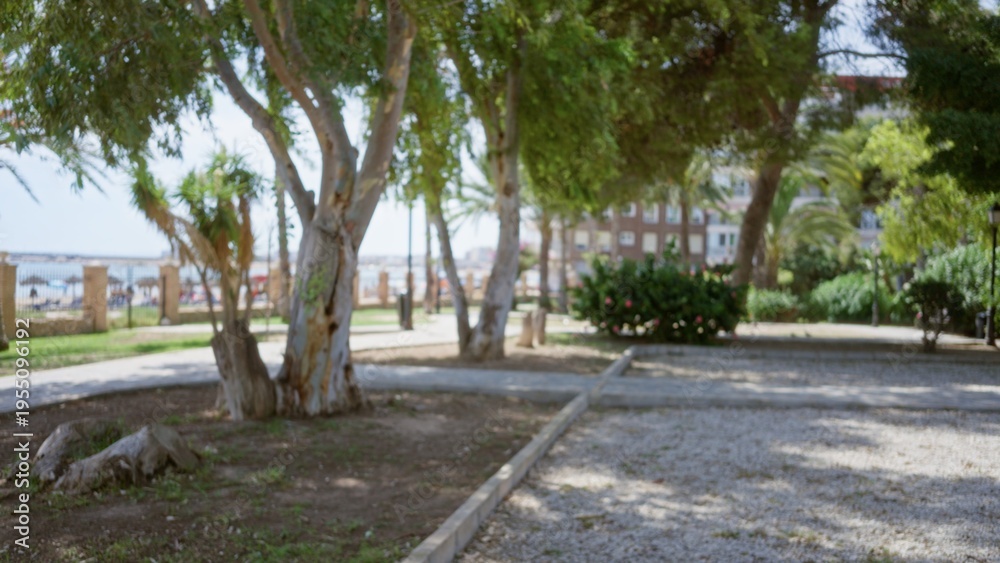 Fototapeta premium Blurred park with soft bokeh trees, defocused promenade and gravel path, shallow focus on foreground trunks; background backplate calm.