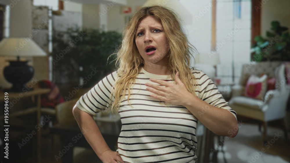 Fototapeta premium Woman clutch chest, mouth open, blurry living room with lamps and armchair, blonde in a striped shirt inside a building; distress.