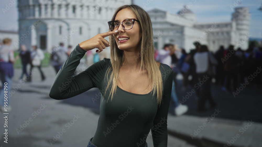 Fototapeta premium Woman points finger to nose at building in pisa wearing glasses and smiling among a crowd; playful travel moment.