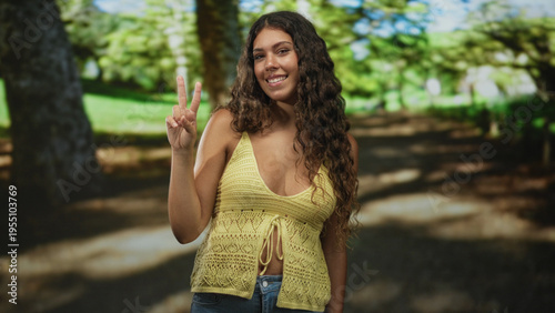Hispanic teenage woman pointing index finger, bare shoulder visible, wearing yellow knit top on forest path; youthful joy.