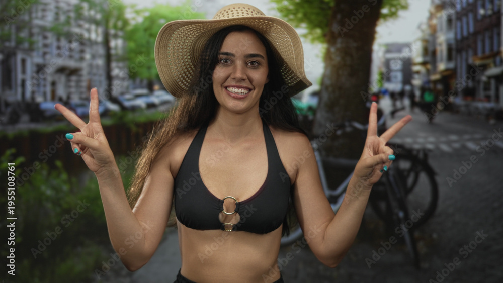 Fototapeta premium Woman holds peace sign and adjusts straw hat on a sunlit tree-lined street near canal and bicycles; urban summer happiness.