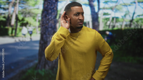 Man in yellow sweater cupping his right ear with hand, grimacing and turning head toward passing joggers on a tree lined path in street; annoyance listening.