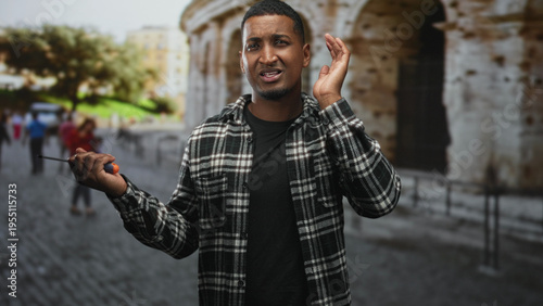 Photos Young black man holding screwdriver and cupping ear on a cobblestone street near an ancient stone facade; confusion seeking help