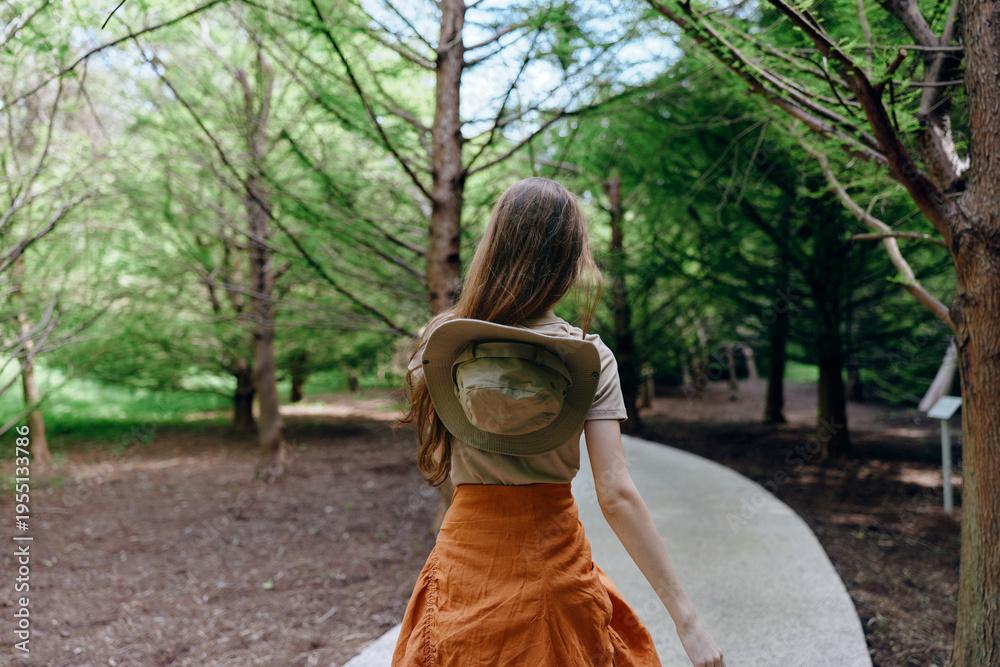 Fototapeta premium Woman walking alone in park among tall trees on a winding path, wearing a hat and orange skirt, back view of solo traveler enjoying nature and peaceful green woodland.