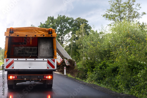 Принти на полотні Roadwork truck warning lights drives along wet asphalt road near green vegetation countryside landscape rain
