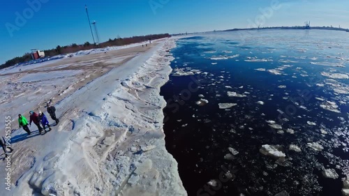 Aerial drone track follows jagged ice and icy berm by sandy snowy beach near the Daugava River mouth in Riga, with small lighthouse and distant port cranes visible.