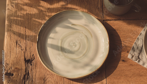 Empty handcrafted ceramic plate on a textured wooden table, softly lit by natural sunlight, casting long, warm shadows
