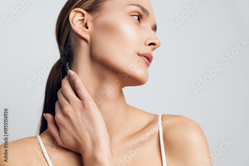 Young woman with smooth skin and natural makeup touches her neck gently, looking sideways with calm expression against a plain light background, showcasing beauty and skincare concept.