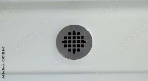 Fotografie Close-up of a circular stainless steel shower drain grate with square holes, set against a pristine white bathtub or shower base, showing minor rust and discoloration around the edges