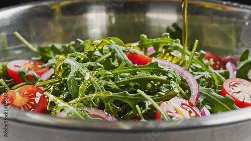 Close-up of olive oil being poured over fresh arugula salad with cherry tomatoes and red onions in a stainless steel bowl
