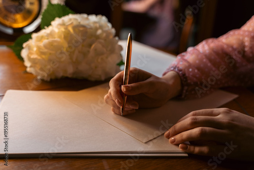 Woman's hands writing a letter with an elegant pen on a vintage wooden desk with flowers.