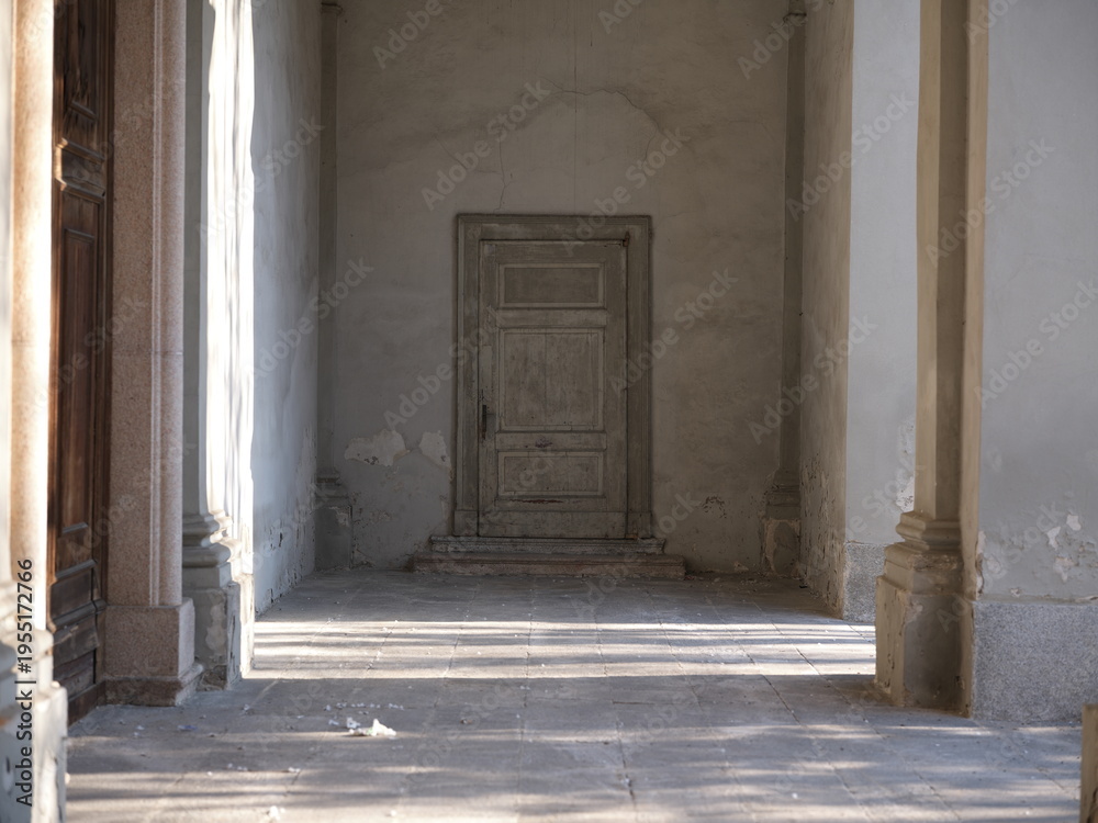 Obraz premium Aged wooden door set in a worn plaster wall with peeling paint. Minimal composition with soft natural light, highlighting texture, decay and timeless rustic character.