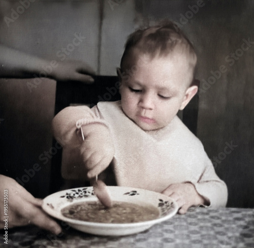 A vintage portrait of a little boy eating from a plate with a spoon. Retro photo from the 1960s.