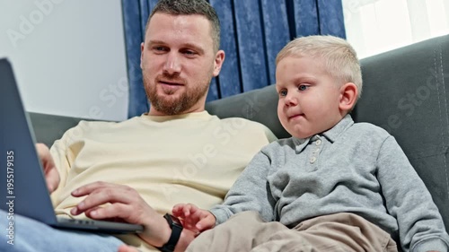 A father is engaged with his laptop on a couch, while his young son expresses displeasure beside him. The light from a nearby window brightens the room.