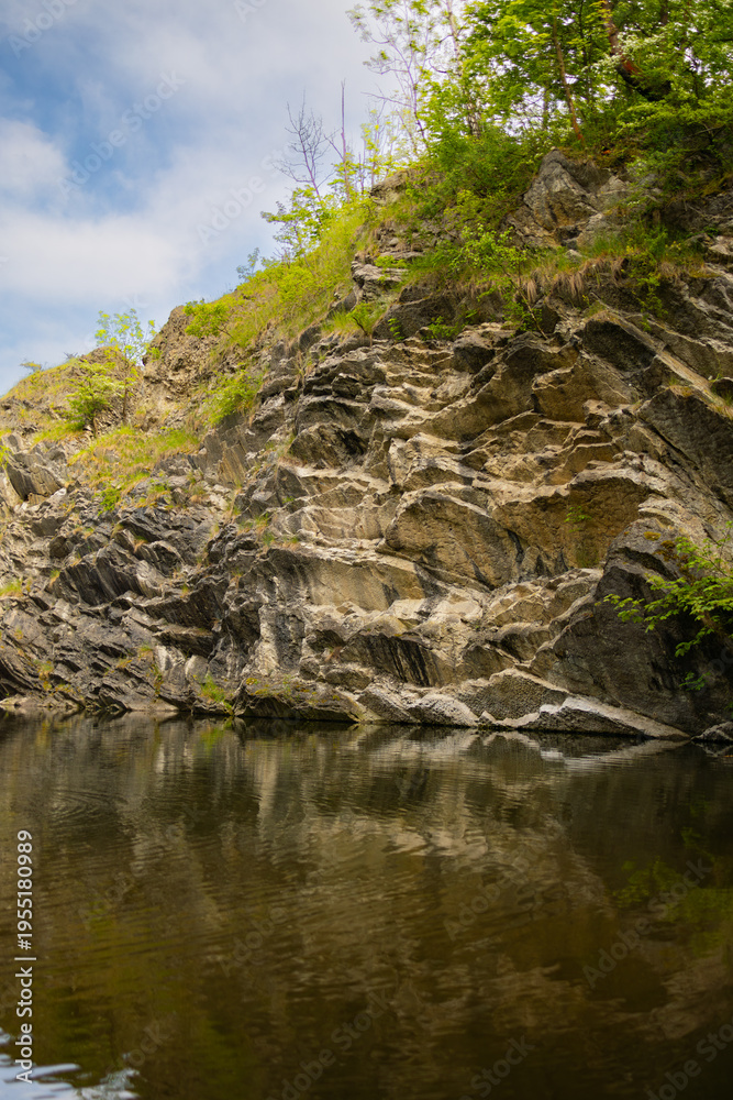 Fototapeta premium Rocky cliffside with lush greenery reflecting in calm water under a partly cloudy sky in a natural outdoor setting