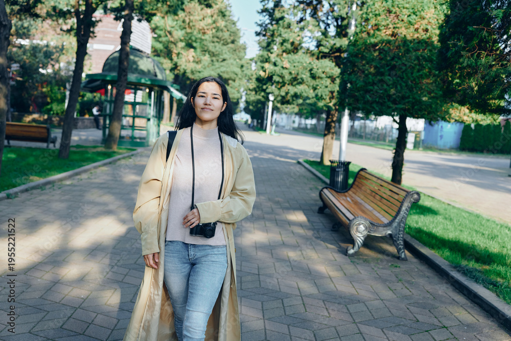 Fototapeta premium Woman walking along a park path with a camera hanging from her neck, wearing a light trench and jeans, enjoying outdoor leisure among trees and benches