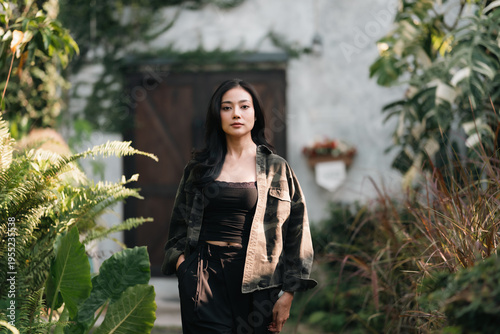 Young woman black hair casual jacket outdoor garden greenery natural light smiling relaxed confident standing foliage nature lifestyle portrait happiness beauty fashionable trendy serene peaceful