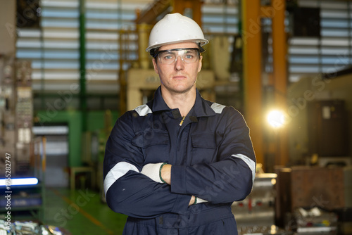 Confident man factory worker in an industrial portrait stands serious with arms crossed wearing safety helmet uniform and glasses feeling professional and proud inside manufacturing plant