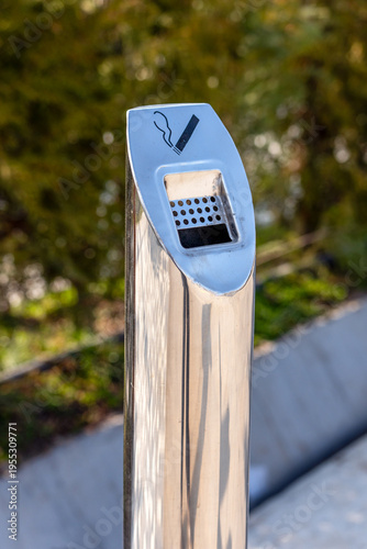 Close-up of a modern, polished stainless steel outdoor ashtray or cigarette disposal bin featuring a clear cigarette and smoke symbol on the top surface.