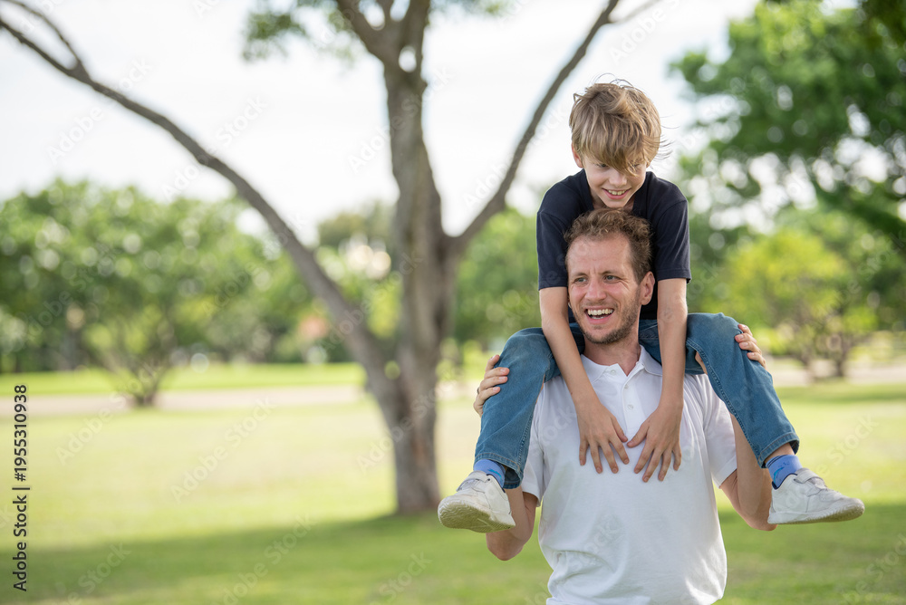 Fototapeta premium Smiling father carrying young son on his shoulders while playing together outdoors in public park. Joyful man giving boy shoulder ride in nature family bonding moment outdoor fun on summer.
