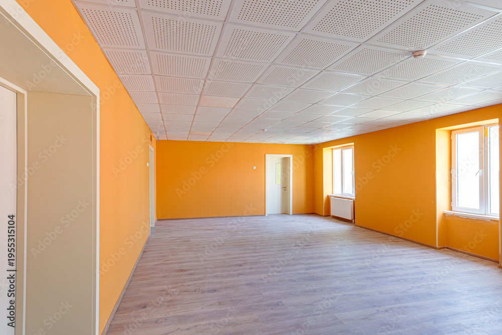 Obraz premium Spacious empty office or classroom interior featuring bright orange walls, wooden laminate flooring, white doors, and a professional acoustic grid ceiling with windows providing natural light.