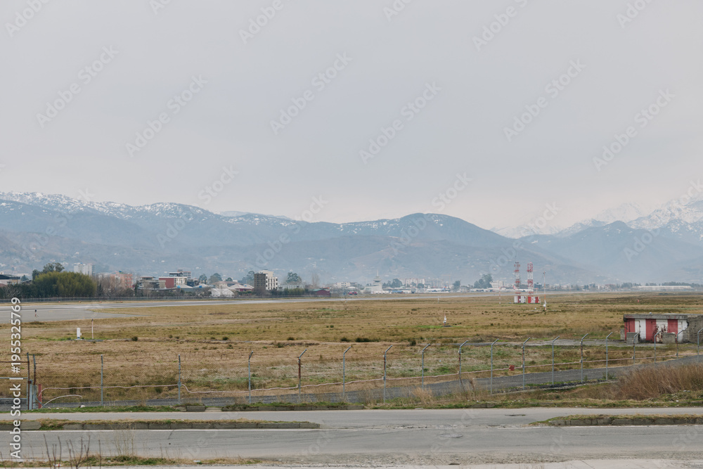 Obraz premium Empty airfield with runway and small buildings in front of hazy mountains under cloudy sky. Concept for aviation, transportation, and industrial landscape with wide open space.