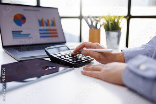 A pair of hands with a tablet and calculator, engaged in financial analysis while surrounded by graphical data in a contemporary office setting, emphasizing modern work dynamics.