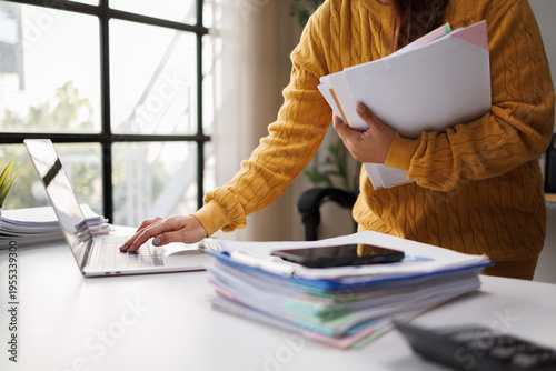 A woman in a yellow knit sweater multitasks at her desk, balancing paperwork and technology, embodying a productive and organized office environment.