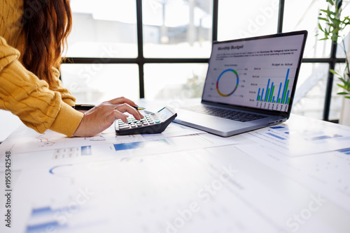 Engaged in budget analysis, a professional uses a calculator and laptop beside financial reports, emphasizing the importance of data accuracy and strategic planning in business contexts.