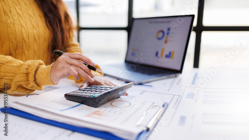 A woman in a cozy yellow sweater is engaged in calculating figures on a calculator while reviewing financial reports next to a laptop, embodying financial diligence.