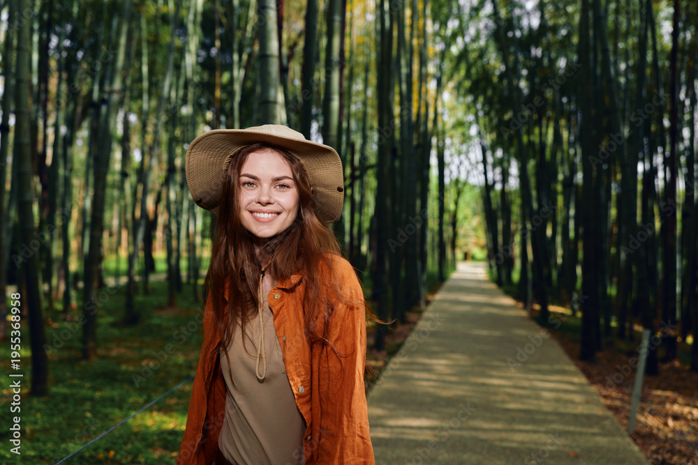 Fototapeta premium Woman smiling in a straw hat walking in an autumn park on a tree lined path, portrait of a happy young adult wearing a cozy jacket with natural light and seasonal foliage.