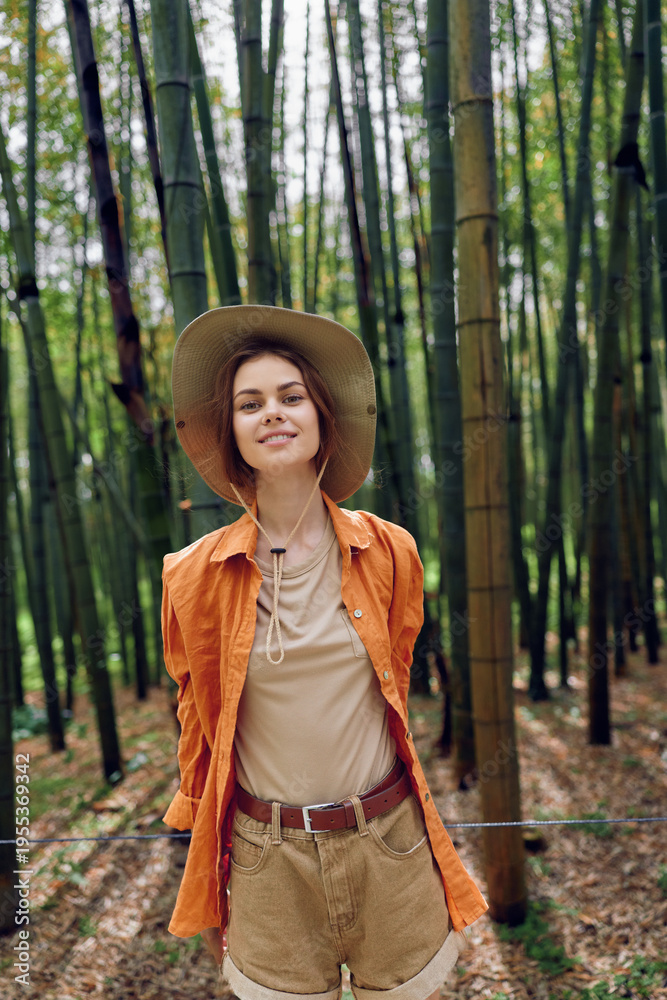 Fototapeta premium Woman in bamboo forest wearing straw hat and casual orange shirt, smiling portrait amid tall green stalks, nature walk in summer outfit, relaxed pose and soft natural light.