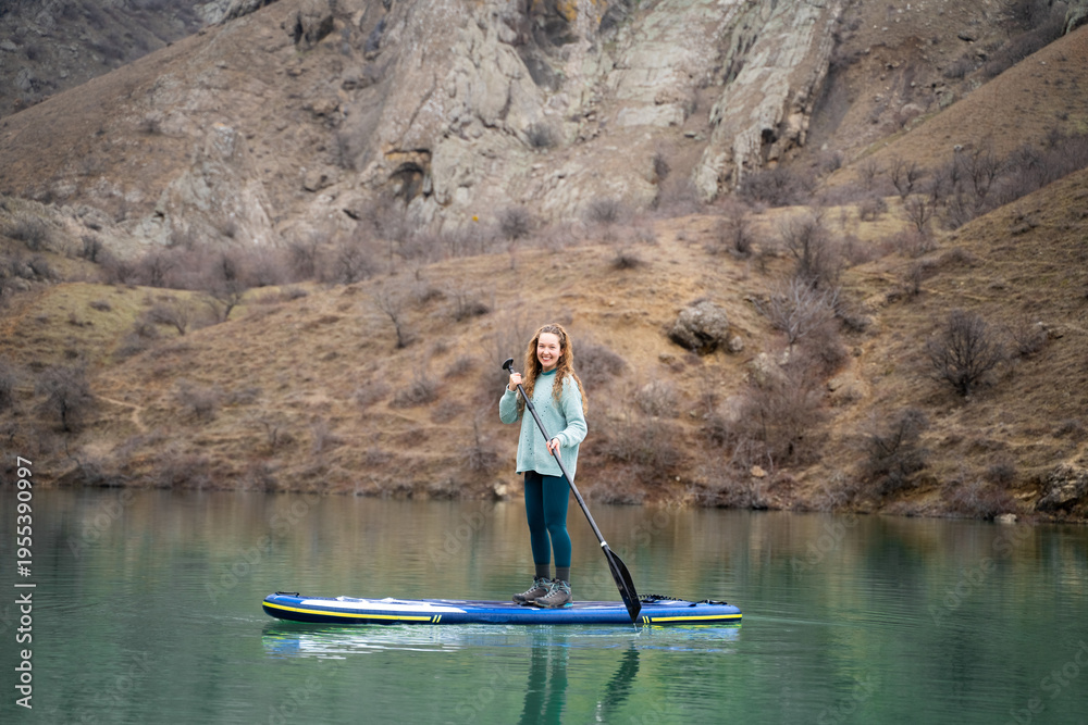 Naklejka premium Woman on paddle board on mountain lake in daylight. Young sporty female on SUP surf on water with mountains in the background. Wide scenic view.