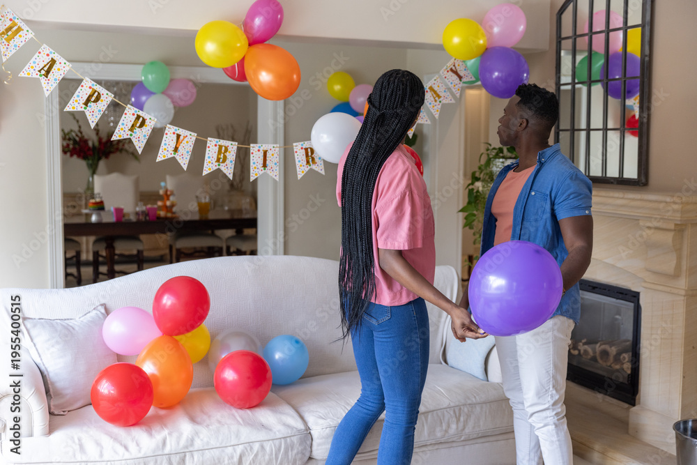 Naklejka premium Couple holding colorful balloons and standing in living room under Happy Birthday banner