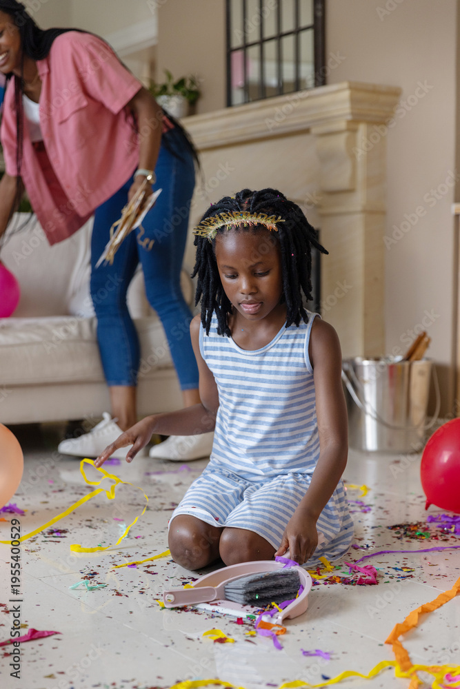 Naklejka premium African American mom and child kneeling, picking confetti with dustpan in living room, balloons