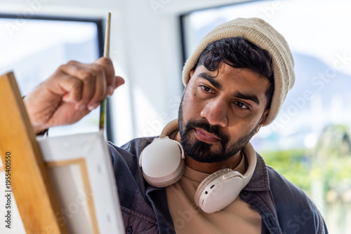 Bild auf Leinwand Indian man painting on canvas on easel in studio holding brush, wearing beanie a