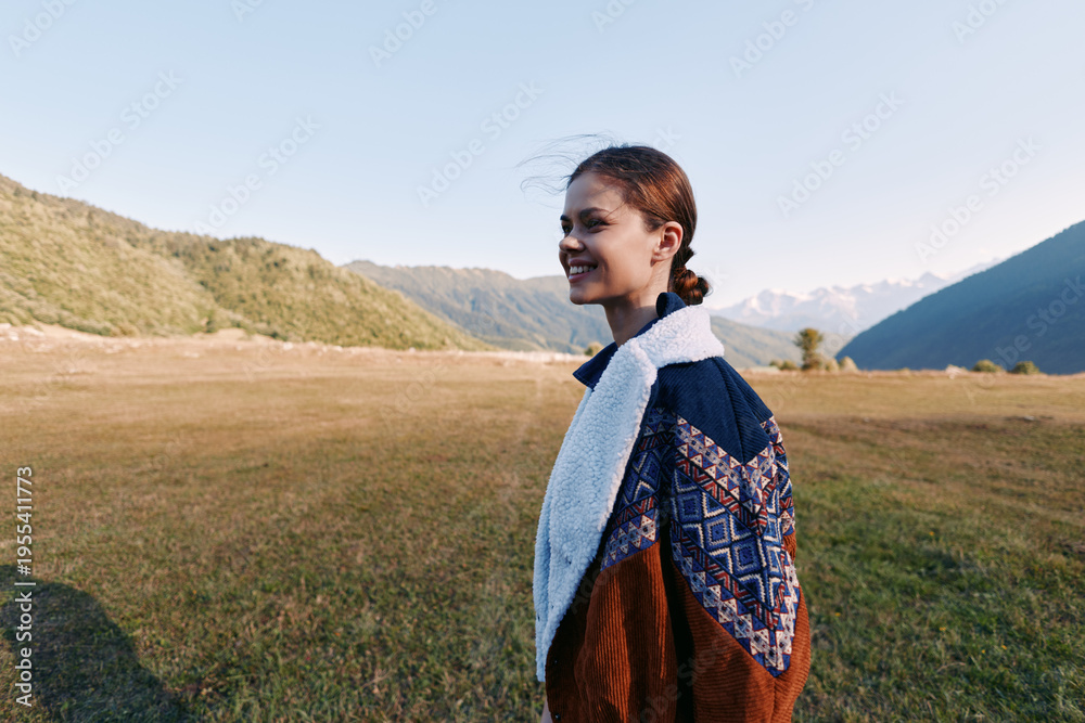 Fototapeta premium Woman, smile, landscape, mountains, meadow, sweater, outdoors, portrait — Young woman smiling in a cozy sweater standing in a meadow by mountain landscape at golden hour for travel and nature.