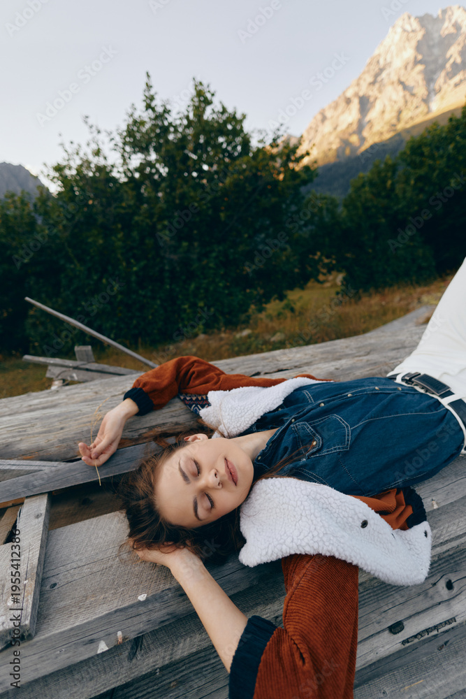 Fototapeta premium Woman portrait relaxing on wooden deck in nature with mountains in background, eyes closed and cozy layered denim and shearling jacket, peaceful outdoor leisure moment and soft evening light