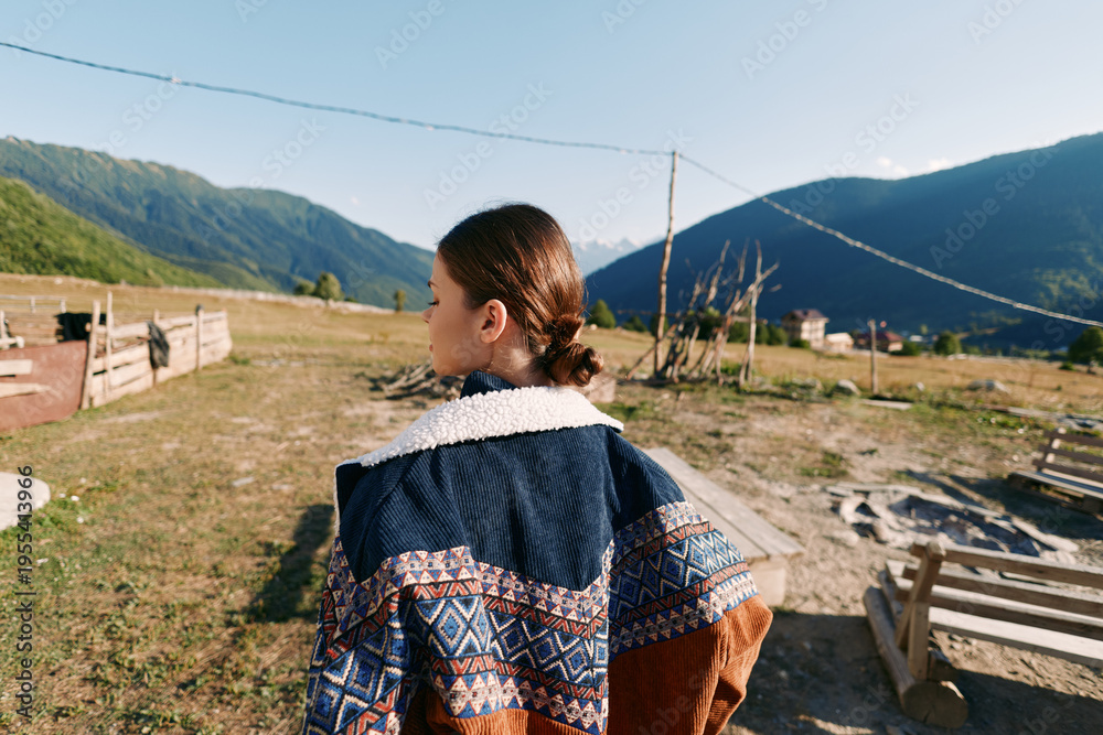 Fototapeta premium Girl in patterned sweater back view in rural village landscape with wooden fences and distant mountains, outdoor portrait of youth exploring countryside pathway under clear blue sunlight.