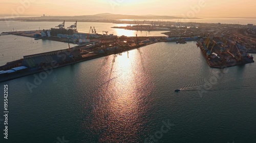 Workers are busy at the port as the sun sets. Small boats move across the water. Cranes and machinery are visible on the shore. The skyline shows buildings and distant hills.