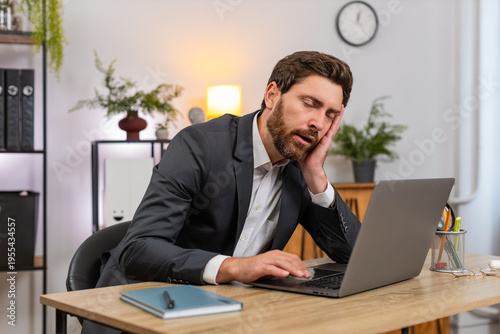 Middle-aged businessman at home office types report on laptop gets sleepy and nods off leaning on hand. Freelancer guy at table fights fatigue blinks slow and almost sleeps then returns to work again.
