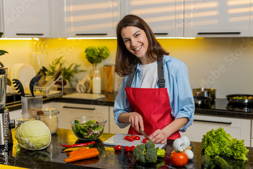 Young woman adds tomatoes seasoning to vegetable salad bowl in home kitchen feeling cheerful meal prep. Adult girl follows healthy diet making fresh lunch for weight loss wellness fit indoors at home.