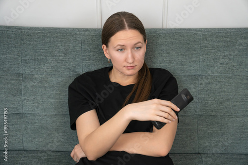 Bored young woman is sits on a comfortable sofa, holding a remote control and switching channels, searching for something interesting to watch on tv