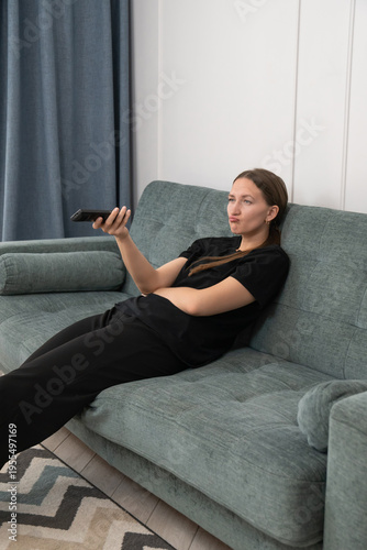 Bored young woman lying on a cozy sofa, holding a remote control while watching television in her living room, indulging in passive leisure and relaxation during the day. Vertical photo