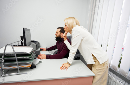 A blonde woman leans over a man seated at a computer, offering guidance in a professional office setting. The scene conveys teamwork, support, and focused collaboration.
