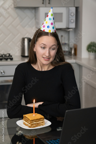 Young woman celebrating her birthday online, wearing a festive party hat and enjoying a delicious piece of cake with a candle, making a wish in her cozy kitchen. Vertical photo