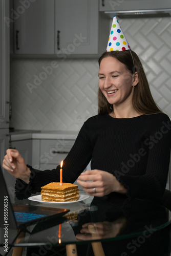 Young woman celebrating her birthday online, wearing a festive party hat while making a wish with a lit candle on a delicious cake, all set up in front of her laptop at home