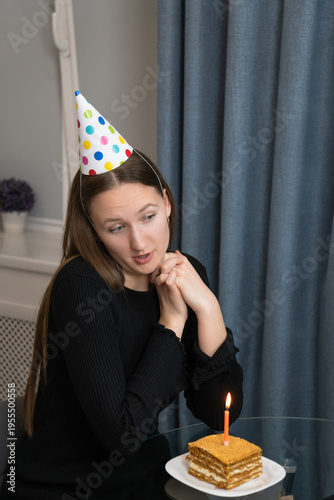 Cheerful young woman wearing a colorful dotted birthday hat smiles and clasps her hands together, making a wish on her special day, sitting in a cozy home environment. Vertical photo